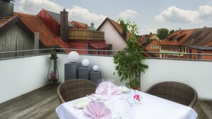 A rooftop terrace with a table set for dining, wicker chairs, and a view of buildings with red roofs. - Alte Post (Wangen im Allgäu)