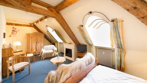 A cozy attic bedroom with a sloped ceiling, wooden beams, a fireplace, a bed, a desk with a lamp, and a television. - Alte Post (Wangen im Allgäu)
