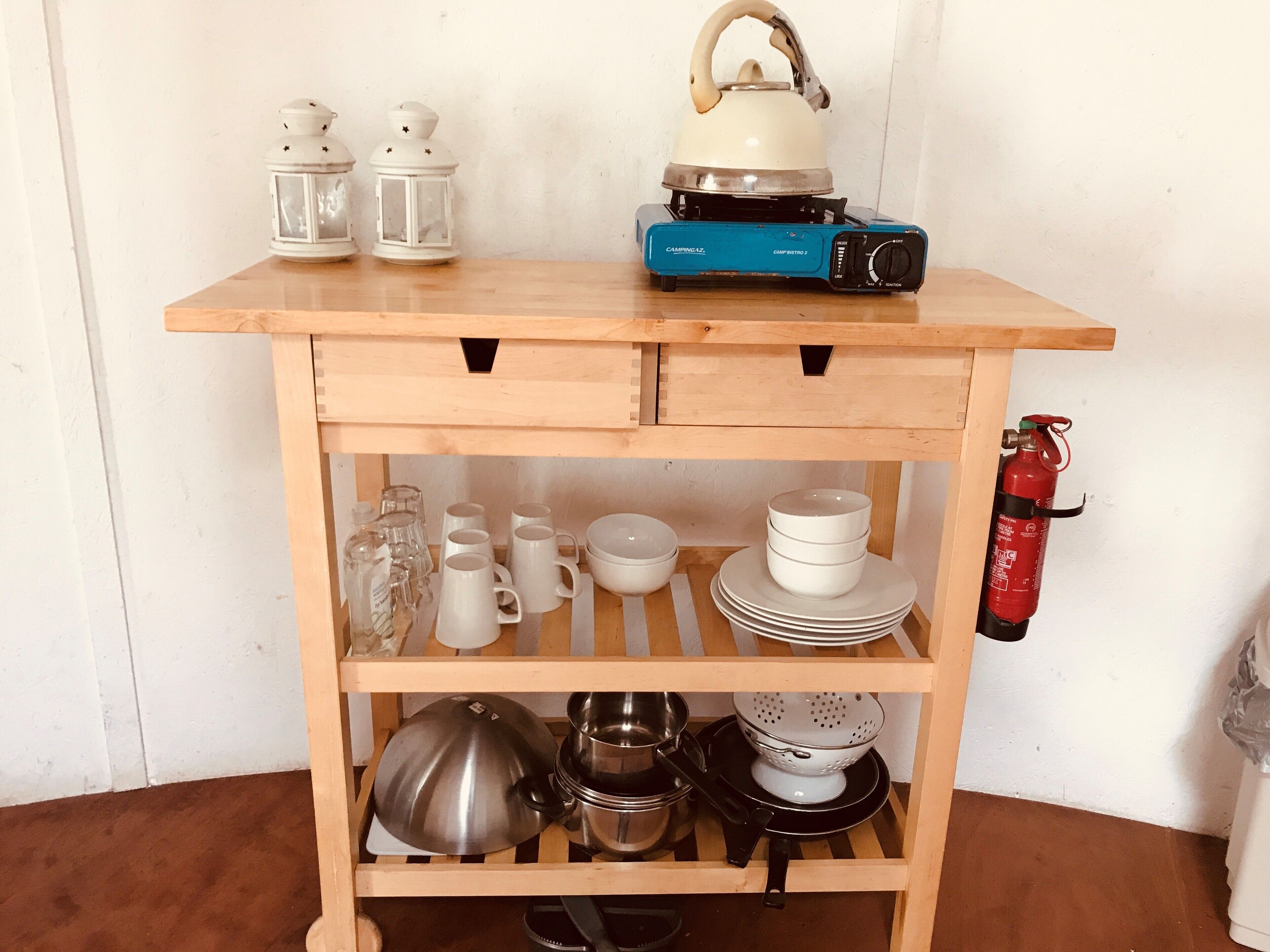 A wooden kitchen cart with shelves holding white mugs, plates, and cookware.