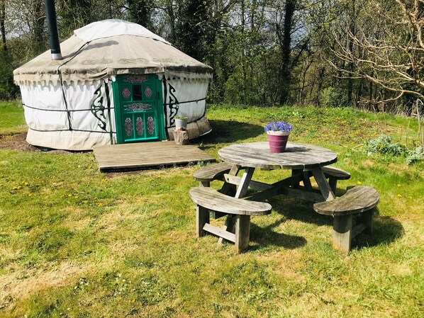 Une yourte avec une porte verte et un petit porche, entourée d’une zone herbeuse où se trouvent une table de pique-nique et des bancs.