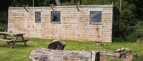 A wooden cabin with three windows, a picnic table, and a log in the foreground.