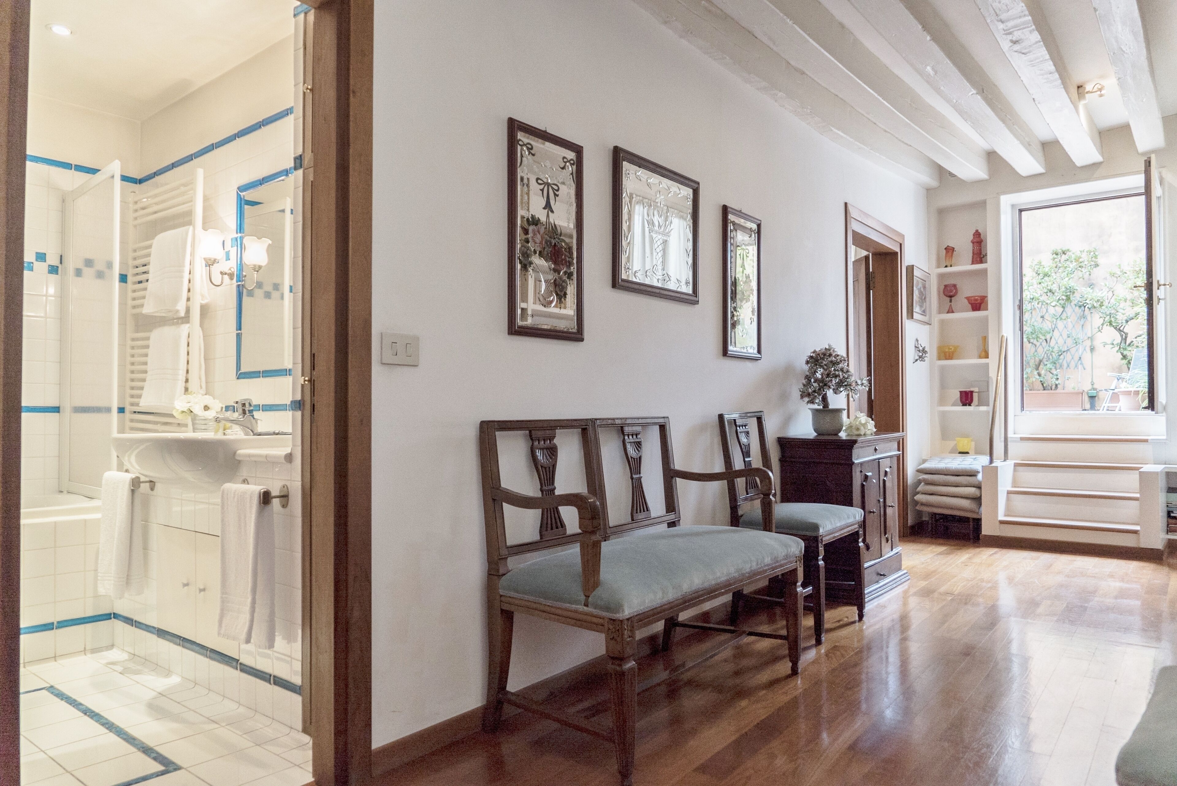 Un pasillo amplio con suelo de madera, un baño blanco con detalles de azulejos y una zona de estar con dos sillas de madera.