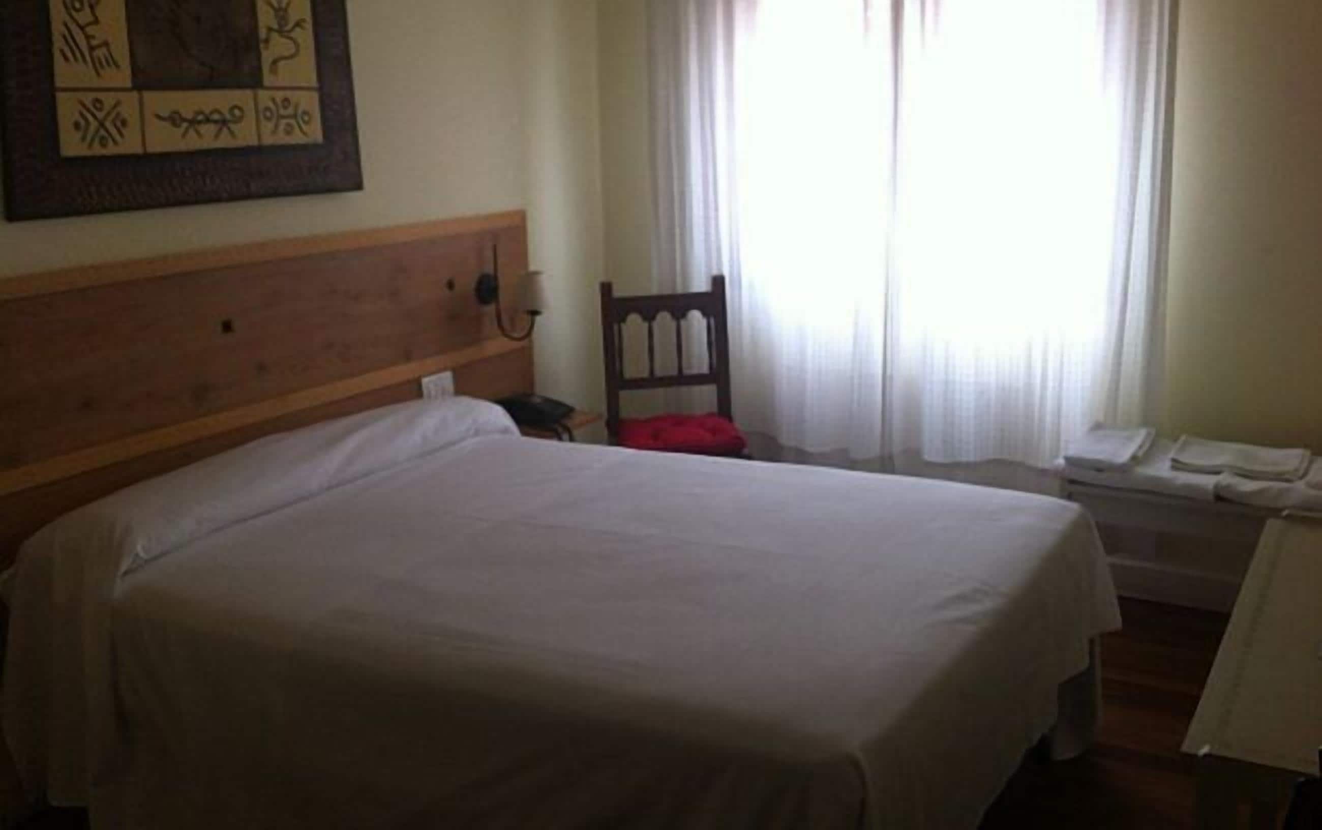A hotel room with a large bed, a wooden headboard, a chair, a small table, and a window with curtains.