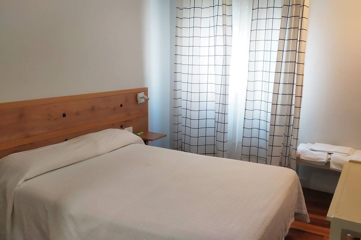 A neatly made bed in a room with a wooden headboard, a small bedside table with a lamp, and a bathroom with a towel rack.