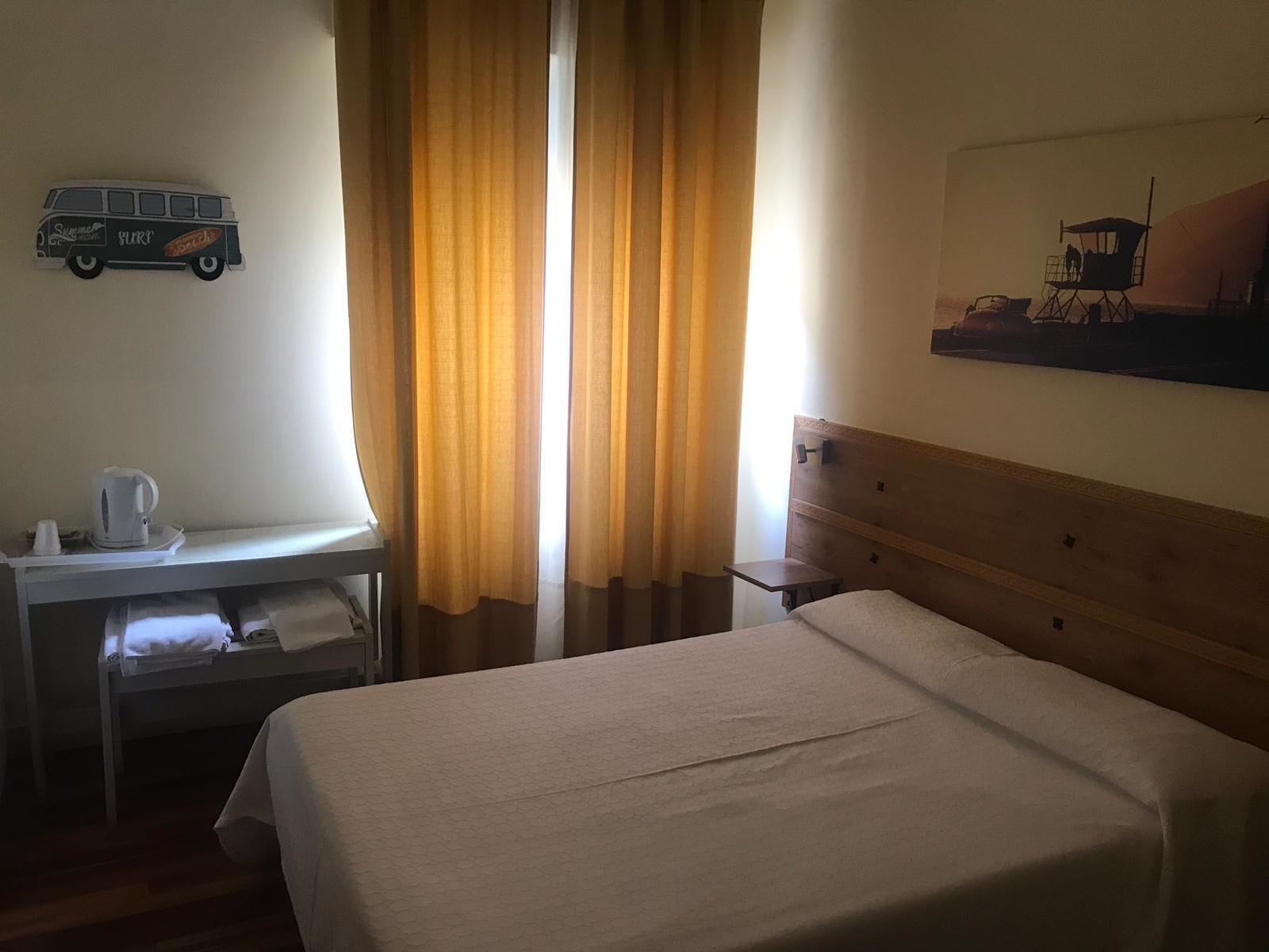 A hotel room with a bed, a desk with a kettle, a chair, and a painting on the wall.