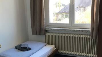 Une petite chambre avec un lit simple, une fenêtre, un radiateur et un lit bien fait, avec une serviette pliée posée dessus.