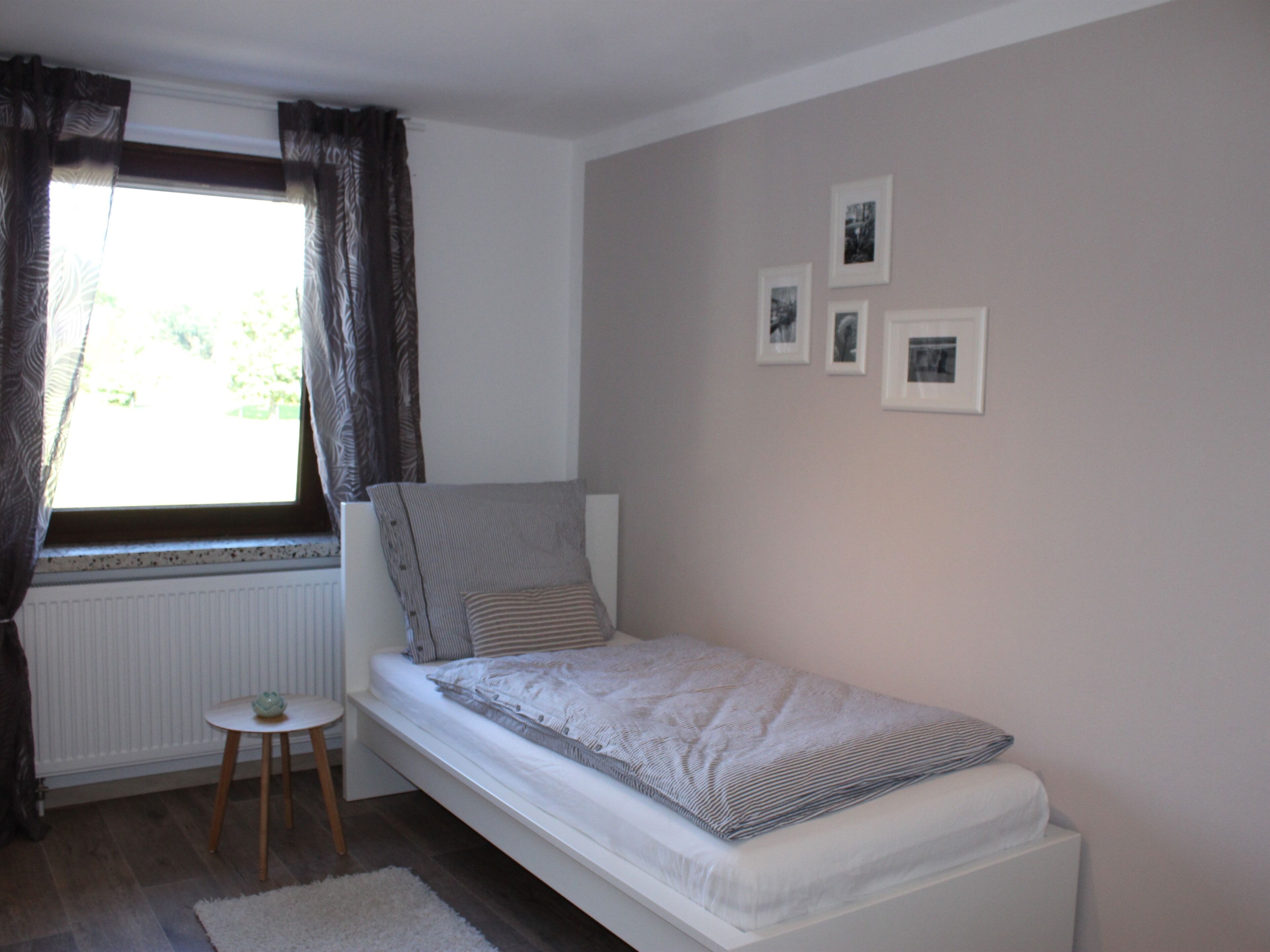 A small, single-bed room with a window, a small table, and three framed pictures on the wall.