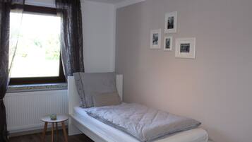 A small, single-bed room with a window, a small table, and three framed pictures on the wall.