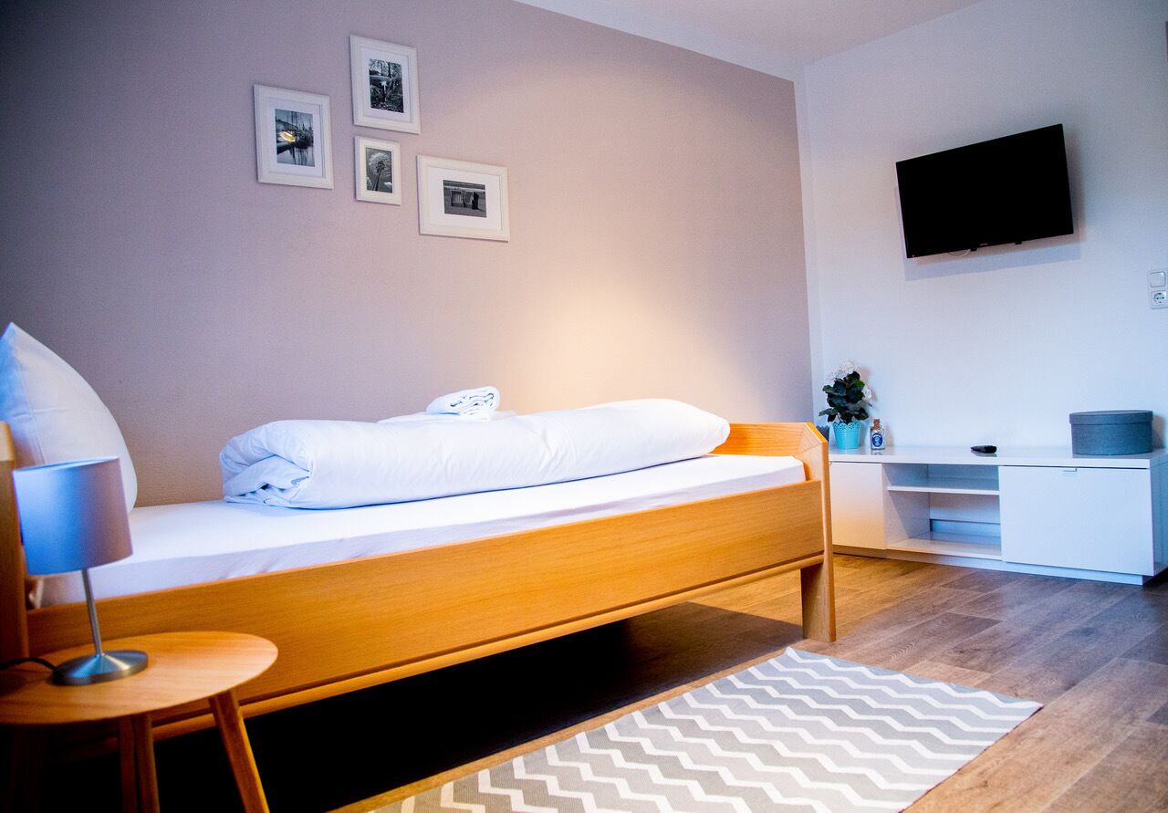 Habitació d'hotel ordenada amb un llit individual, una taula de nit amb una làmpada, un televisor fixat a la paret i una petita zona de treball.