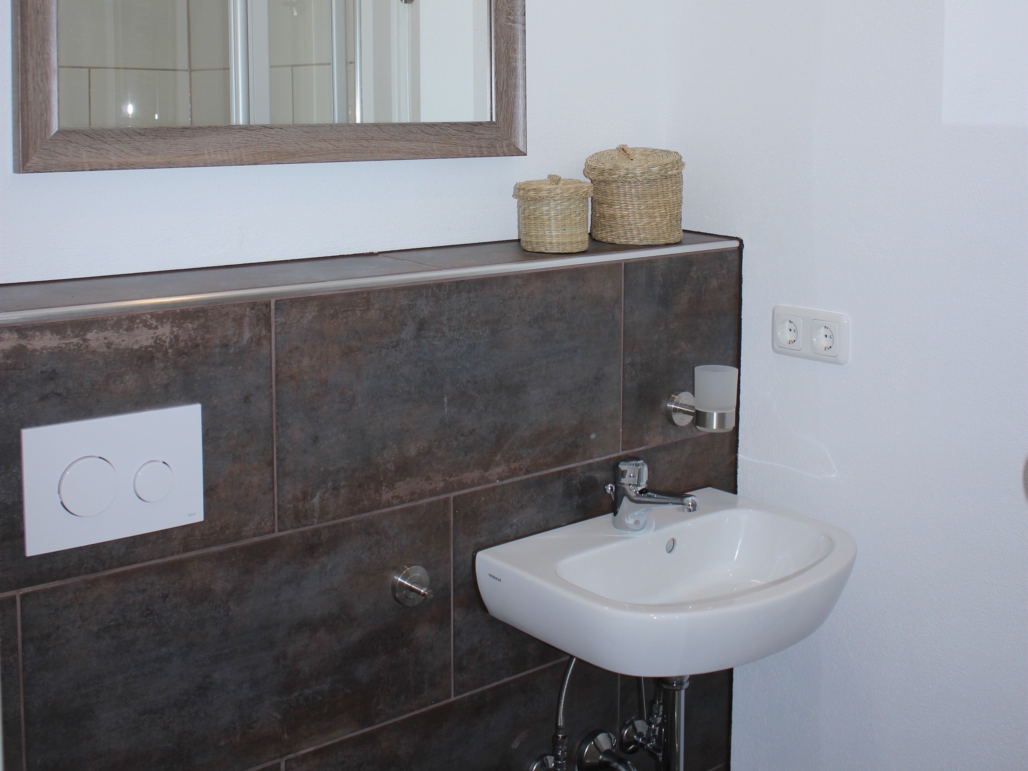 Ein Badezimmer mit einem weißen Waschbecken, dunkelbraunen Fliesen, einem Spiegel und zwei Korbwäschekörben an der Wand.