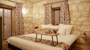 Ein Schlafzimmer mit Steinwand, floralem Vorhang, Bett mit weißer Bettwäsche, Nachttisch mit Getränk und roter Rose sowie einer kleinen Pflanze auf dem Nachttisch.