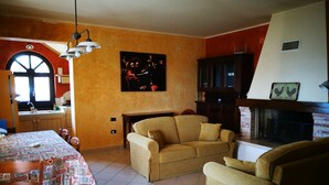A living room with a sofa, a dining table with chairs, a fireplace, and a kitchen area with a window and sink. - Villa Agreste (Cupello)