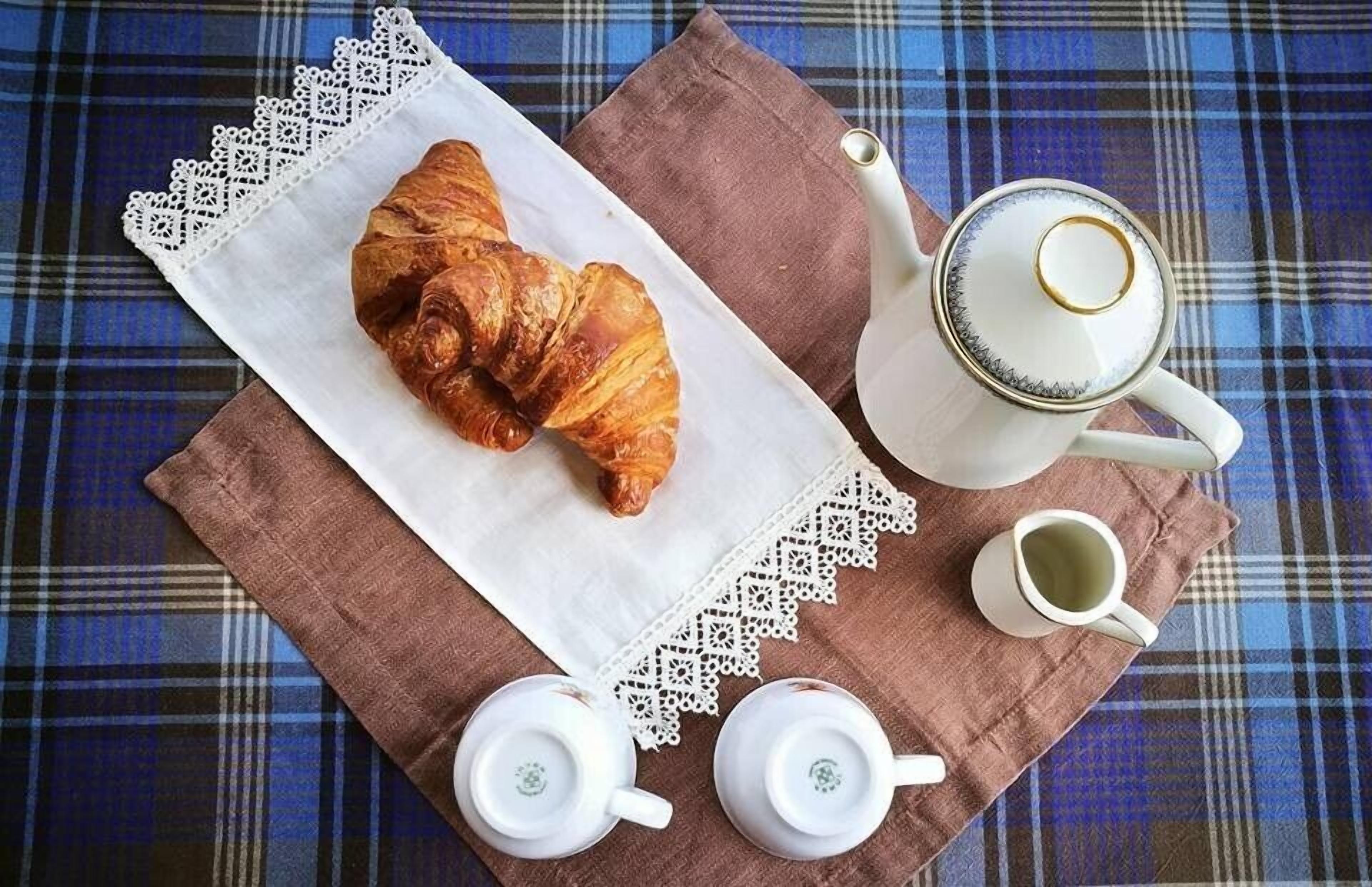 Ein Croissant auf einer Serviette, ein Teekessel und zwei Teetassen auf einer karierten Tischdecke.