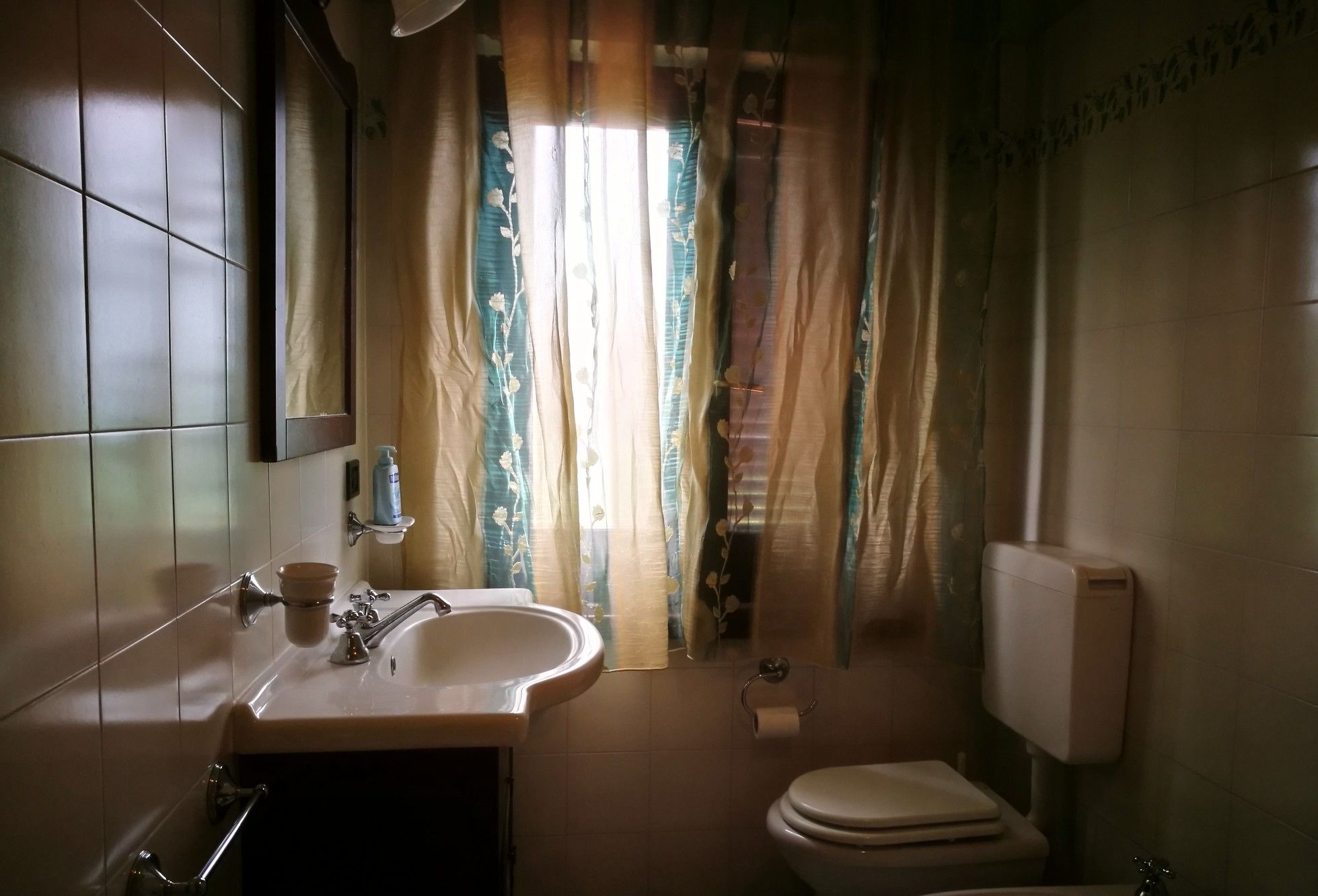 A bathroom with a sink, mirror, toilet, and a window with floral curtains.