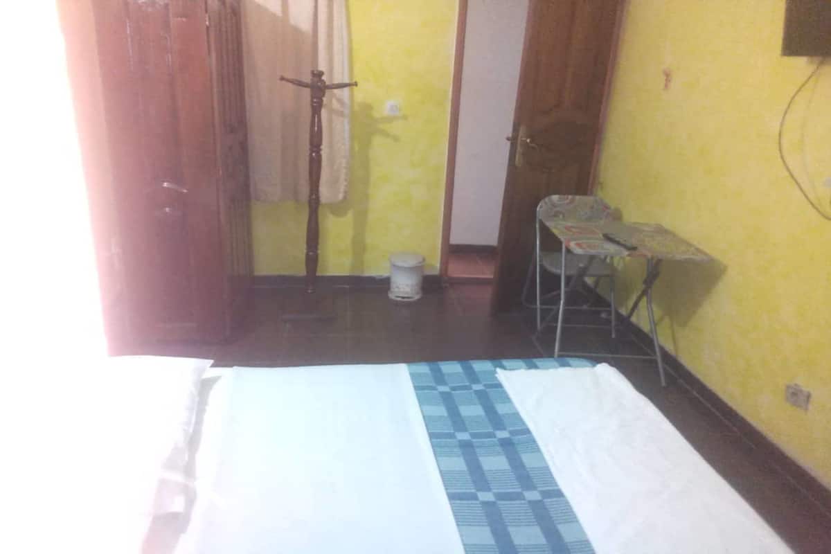 A room with a bed, a small table, and a chair. There is a door leading to another room and a window with curtains.