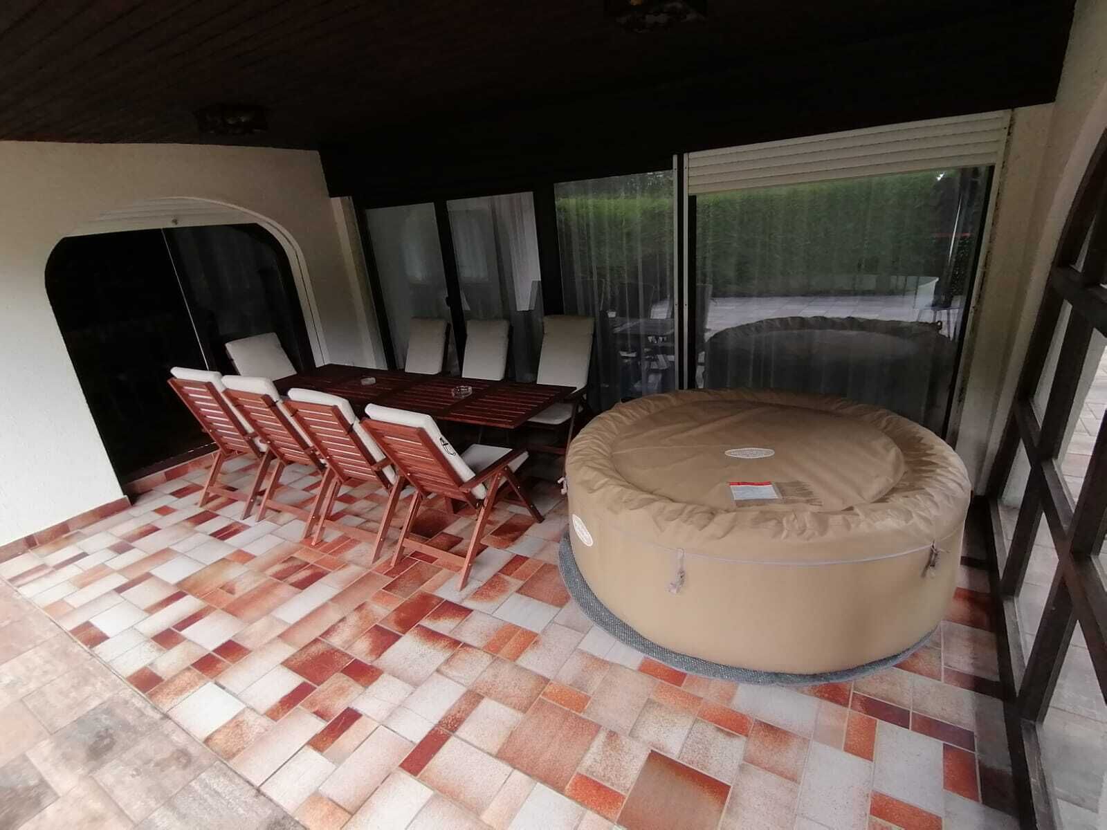 Een overdekt terras met een eettafel en stoelen, een relax fauteuil en uitzicht door grote glazen deuren.