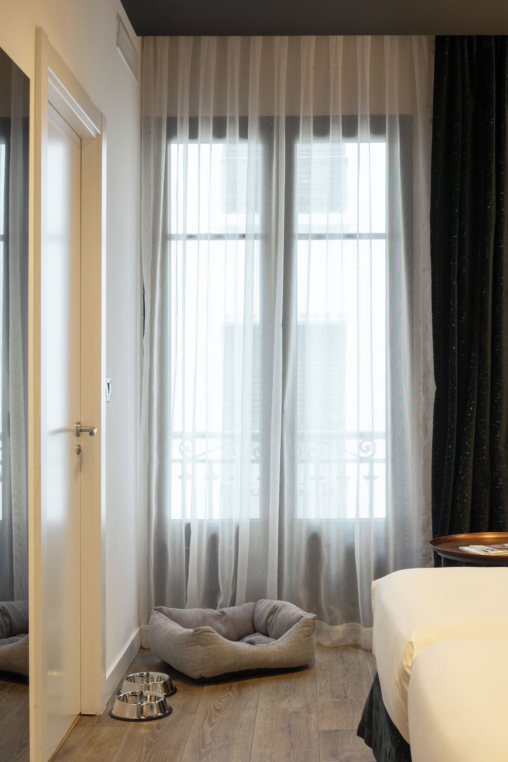 A hotel room with a large window, a pet bed, a bed, and a small table with books.
