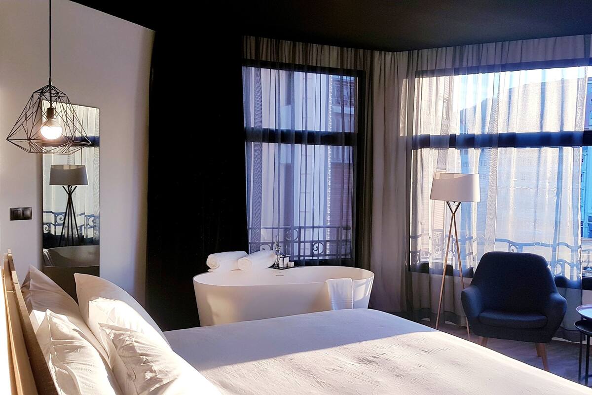 A hotel room with a large bed, a desk, a chair, a lamp, and a view of buildings through the windows.