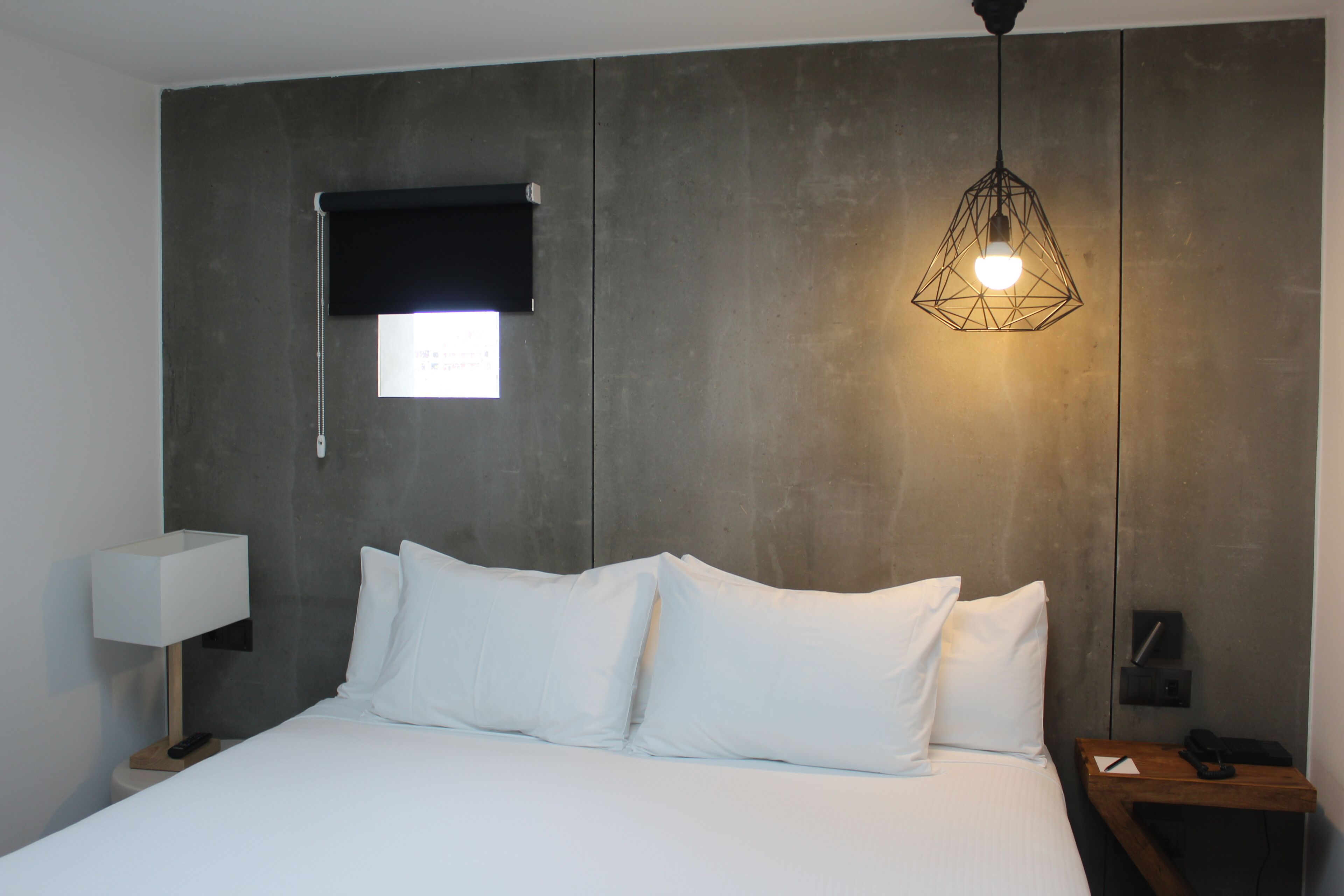 Una habitación de hotel moderna con una cama grande, ropa de cama blanca, una mesita de noche con una lámpara, una lámpara colgante y una ventana pequeña con persiana.