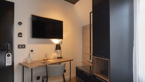 Una habitación moderna y compacta con escritorio de madera, una silla, un televisor de pantalla plana en la pared, una mesita con lámpara y estanterías integradas.