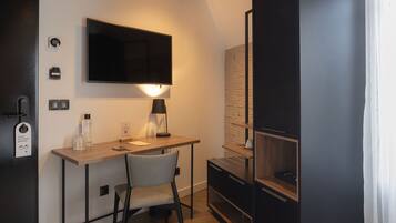 A modern, compact room with a wooden desk, a chair, a flat-screen TV mounted on the wall, a small table with a lamp, and a built-in shelving unit.
