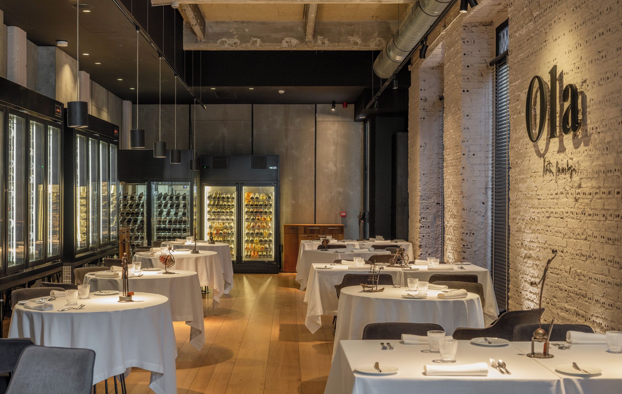 Interior de restaurante con varias mesas, refrigeradores de vino y una pared de ladrillo con el nombre 'Olga'.
