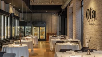 Interior de restaurante con varias mesas, refrigeradores de vino y una pared de ladrillo con el nombre 'Olga'.