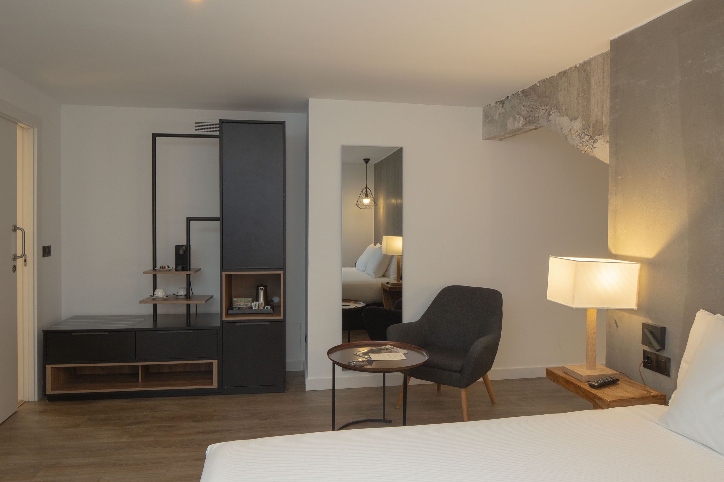 A modern hotel room with a bed, a desk, a chair, a lamp, and a nightstand.