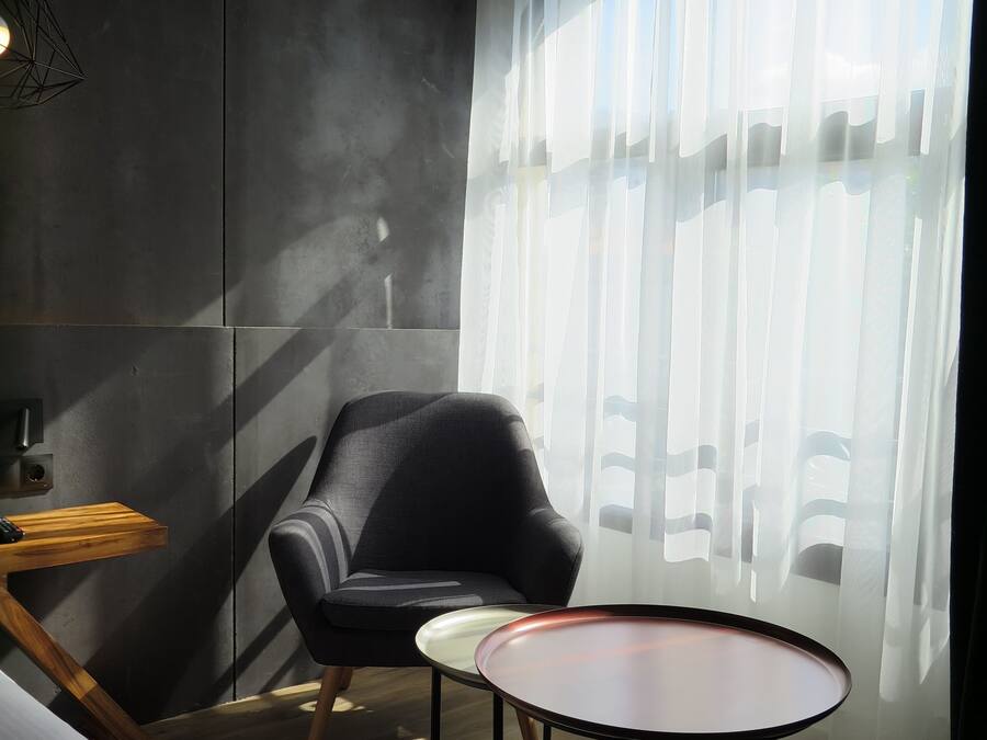 Una habitación moderna con una pared gris oscuro, un sillón negro, una pequeña mesa redonda y una ventana con cortinas de gasa.