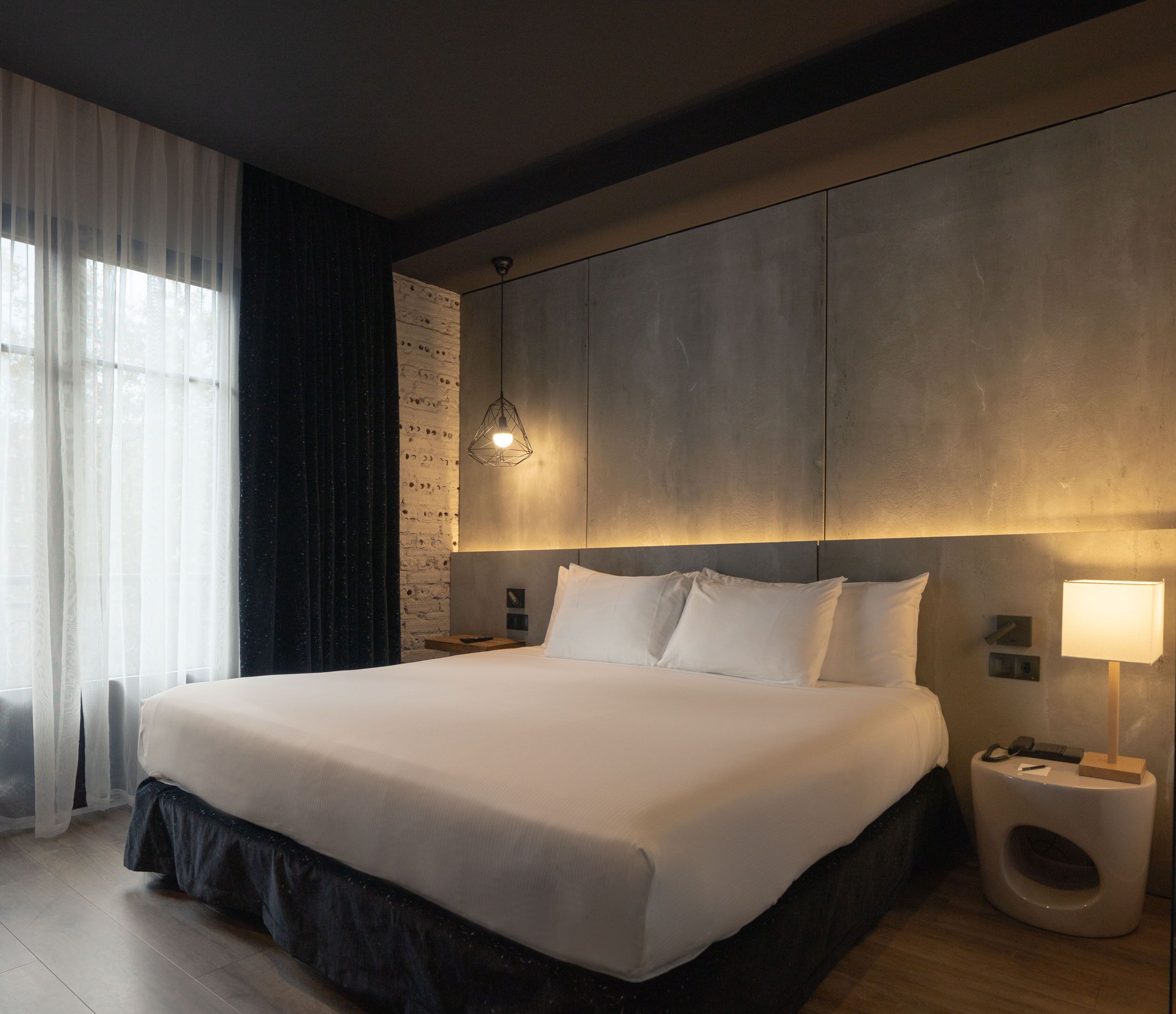 A modern hotel room with a large bed, a bedside table with a lamp, and a wall-mounted light fixture.