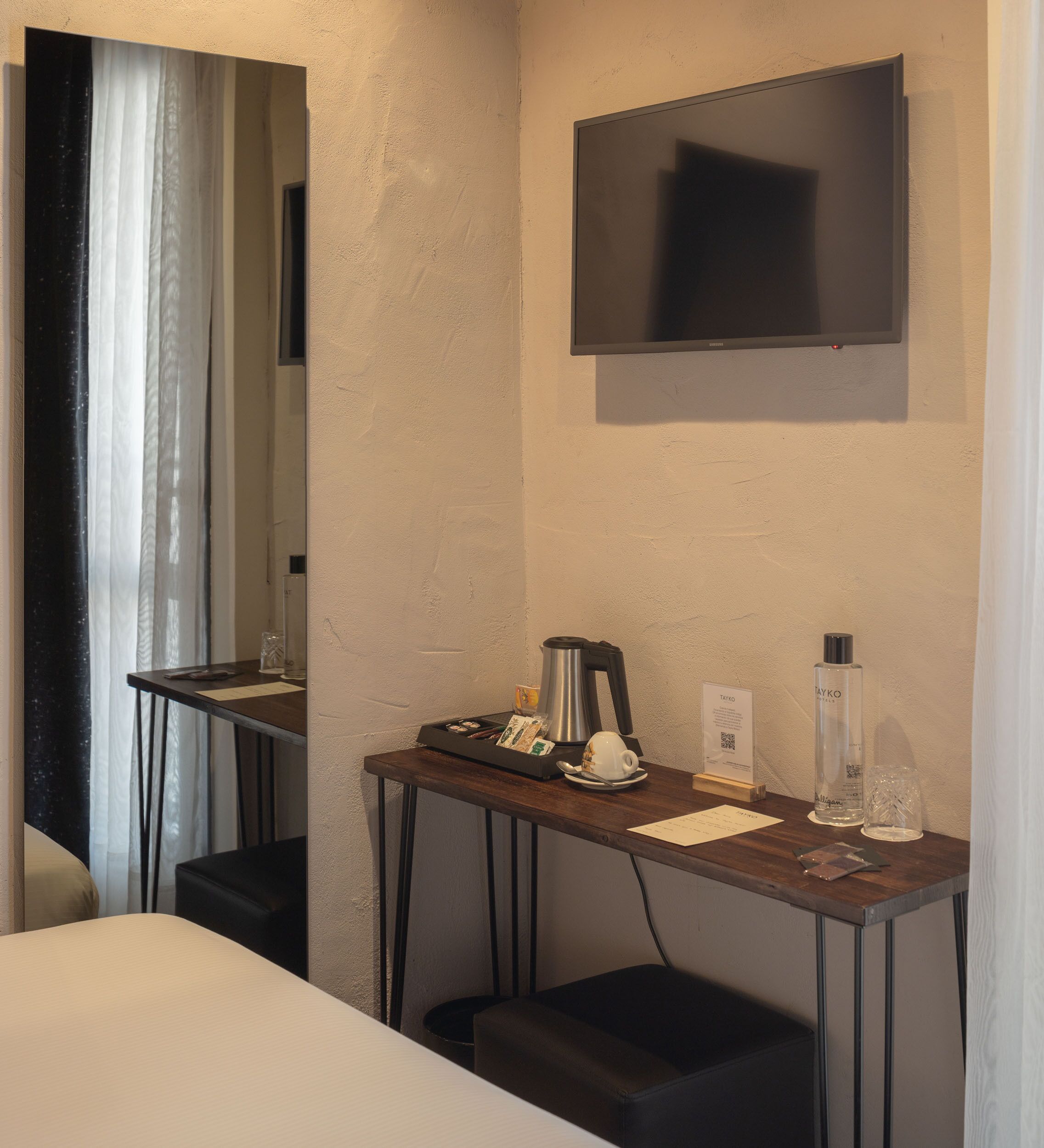 A hotel room with a bed, a desk with a coffee maker, a bottle of hand sanitizer, a TV mounted on the wall, and a mirror.