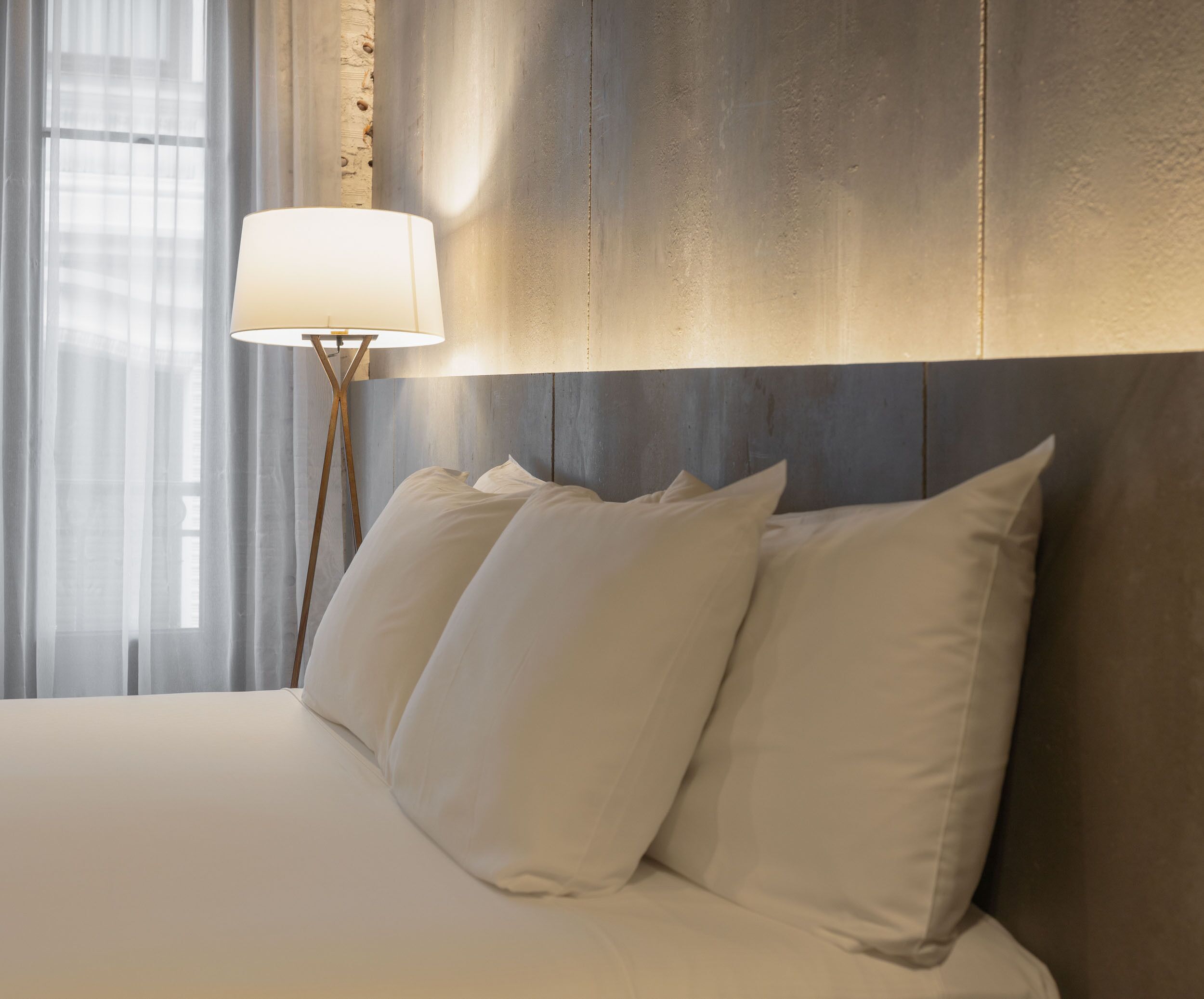 Una cama bien hecha con sábanas y almohadas blancas, una lámpara moderna con base de madera, y un amplio ventanal con cortinas translúcidas.