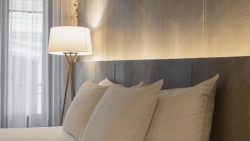 Una cama bien hecha con sábanas y almohadas blancas, una lámpara moderna con base de madera, y un amplio ventanal con cortinas translúcidas.