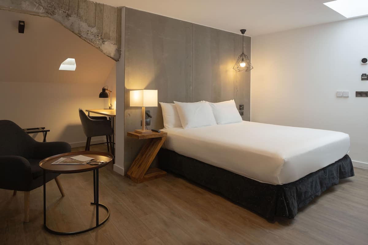 A modern hotel room with a large bed, a desk with a chair, and a small table.