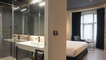 Ett modernt hotellrum med en dusch som är avskärmad med glasväggar, en stor spegel, ett vitt handfat och en säng med en sänggavel i trä.