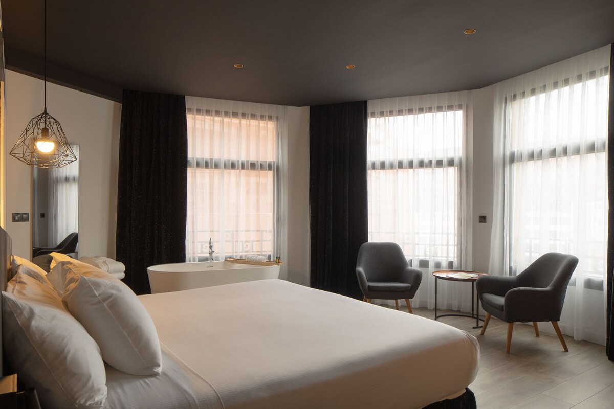 Une chambre d’hôtel moderne avec un grand lit, deux fauteuils, une petite table et un luminaire suspendu.