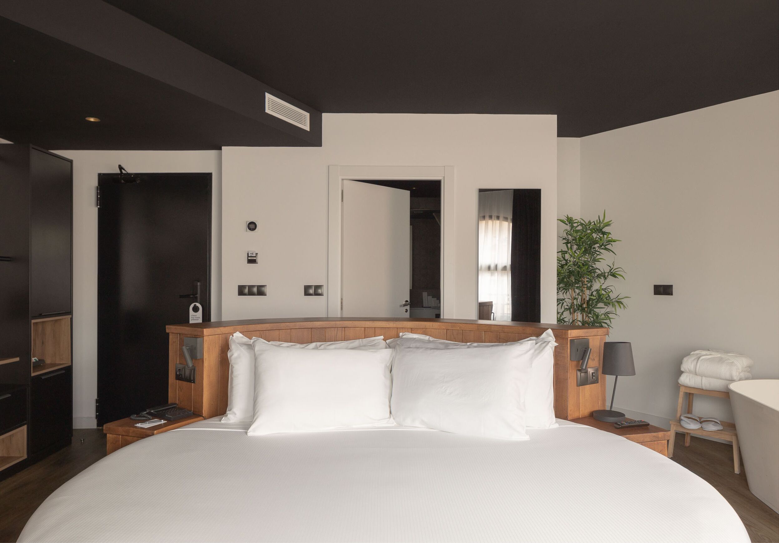Habitación de hotel moderna con una cama grande, cabecera de madera, mesitas de noche, un espejo, una planta y una pequeña zona de estar.