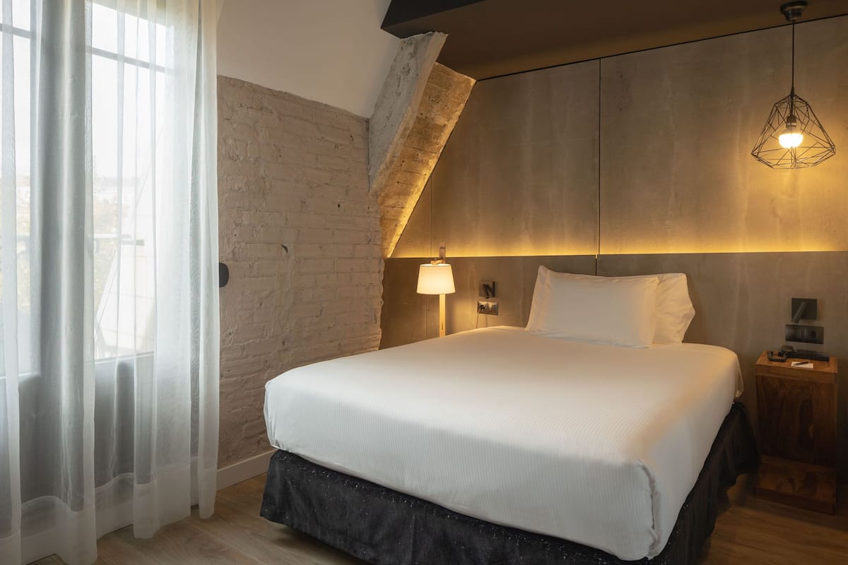 A modern bedroom with a large bed, a wooden headboard, a sloped ceiling, and a hanging light fixture.