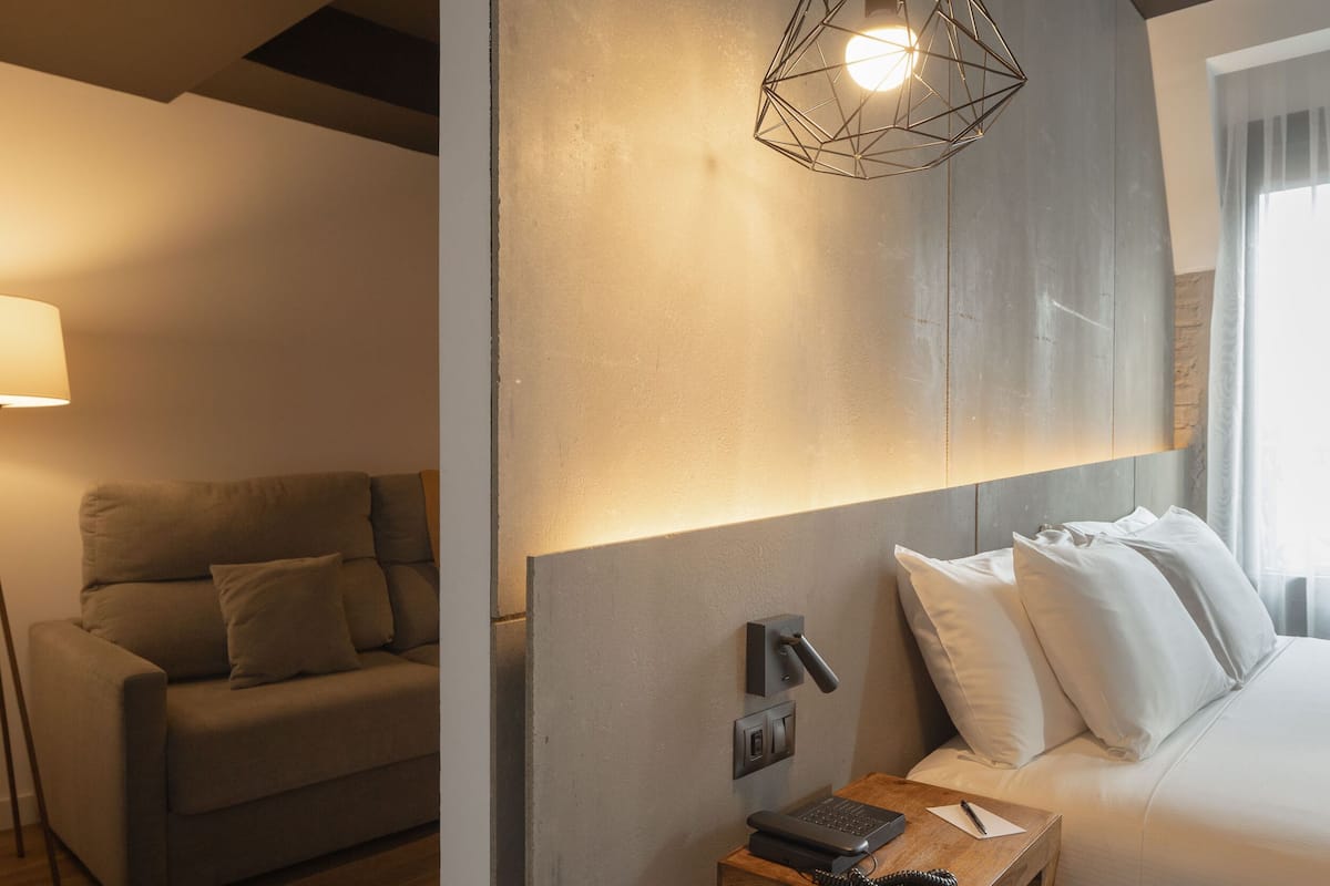 A modern bedroom with a geometric pendant light, a bedside table with a phone and a pen, a sofa, and a bed with white bedding.