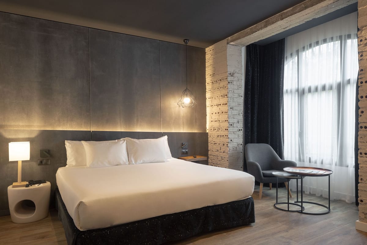 A modern hotel room with a large bed, a bedside table, a lamp, a chair, and a small table with a tray.