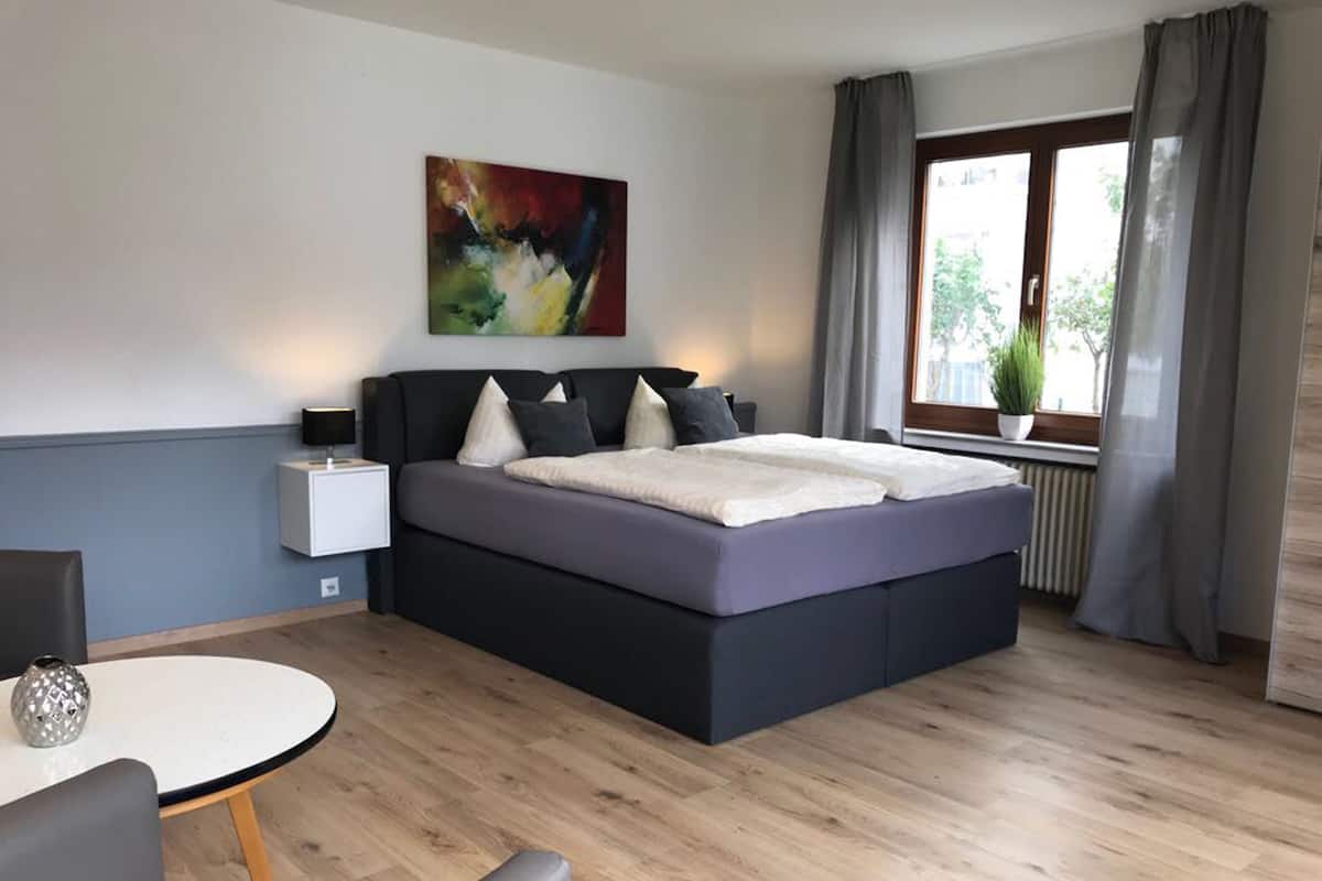 Une chambre à coucher moderne avec un grand lit, une table de chevet, une chaise, une table ronde et une fenêtre avec des rideaux.