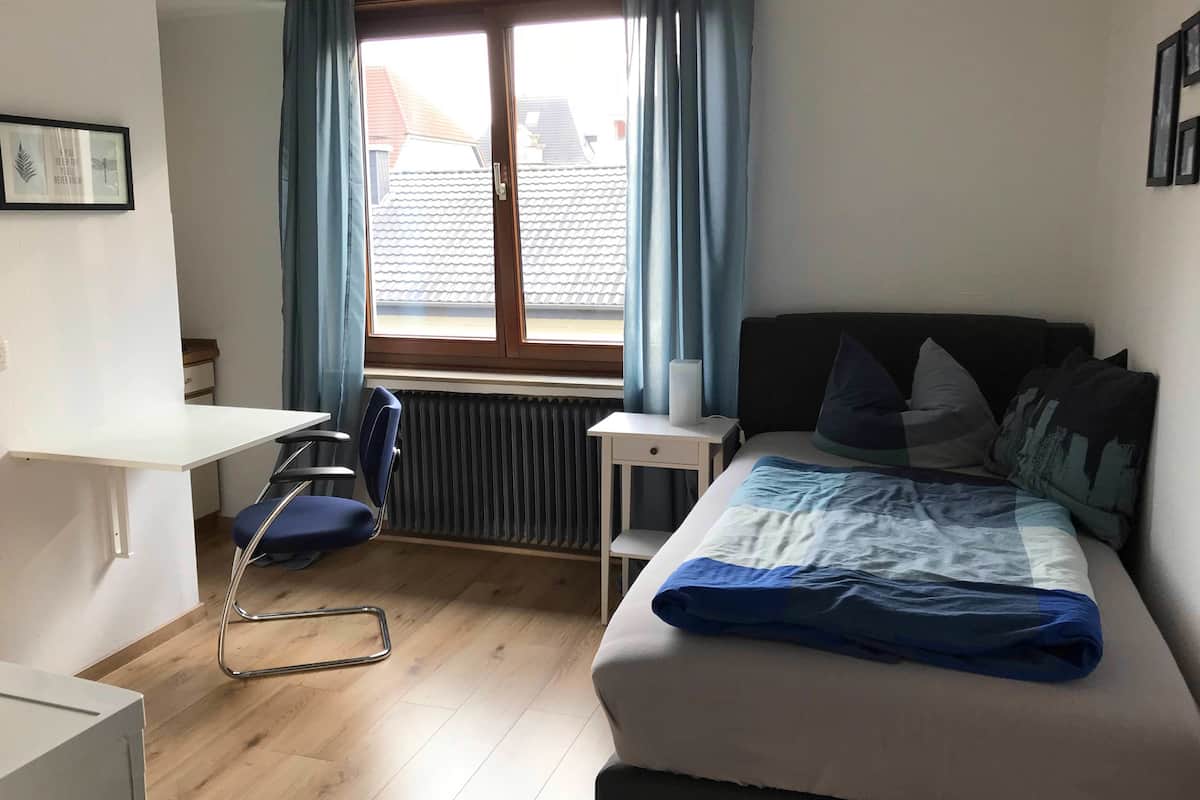 Une chambre à coucher avec un lit, un bureau, une chaise, un radiateur et une fenêtre avec des rideaux.