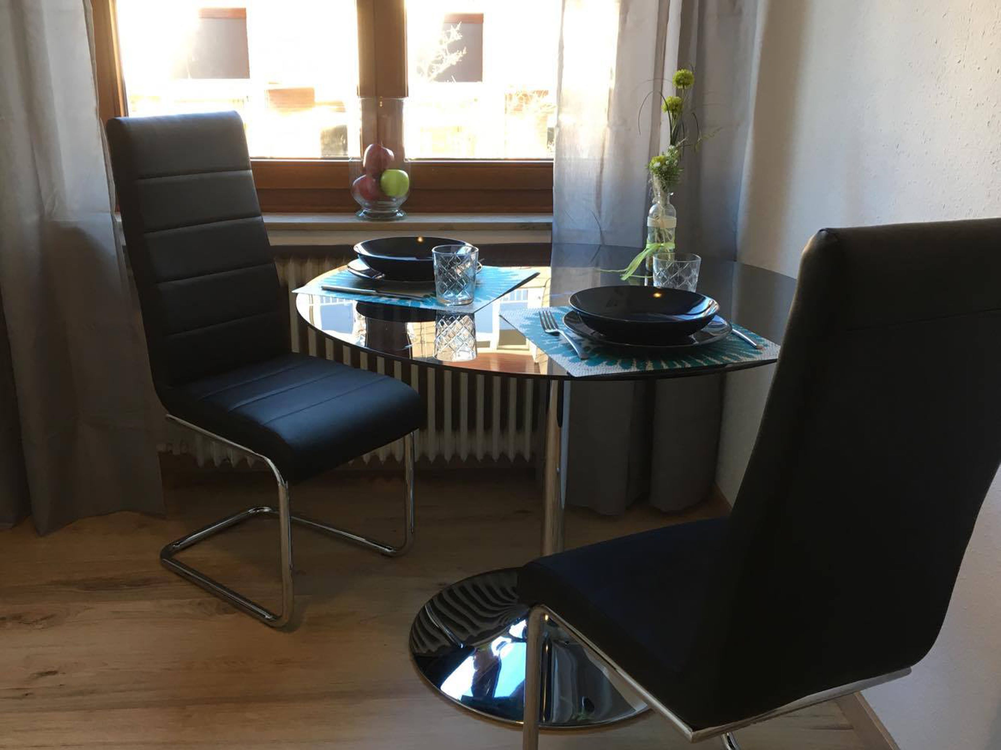 Un comedor con dos sillas negras, una mesa redonda de cristal y un florero con flores en el alféizar de la ventana.