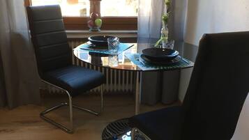 Een eetgedeelte met twee zwarte stoelen, een ronde glazen tafel en een vaas met bloemen op de vensterbank.