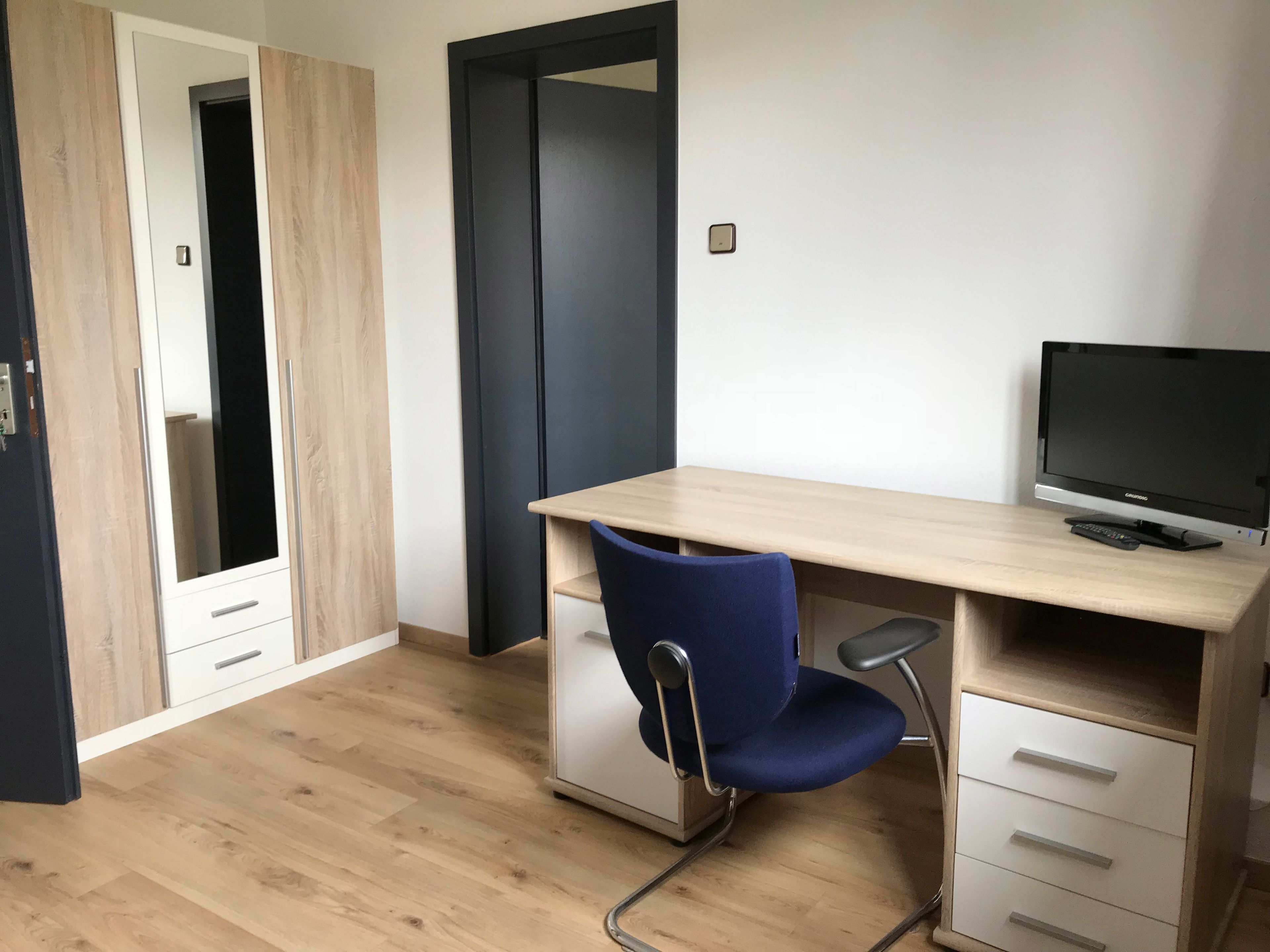 A room with a wooden desk, a blue chair, a TV, and a wardrobe.