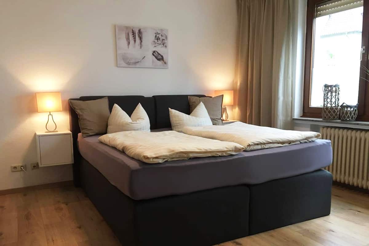 Une chambre moderne avec un grand lit, deux lampes de chevet, une fenêtre avec des rideaux et une œuvre d’art murale.