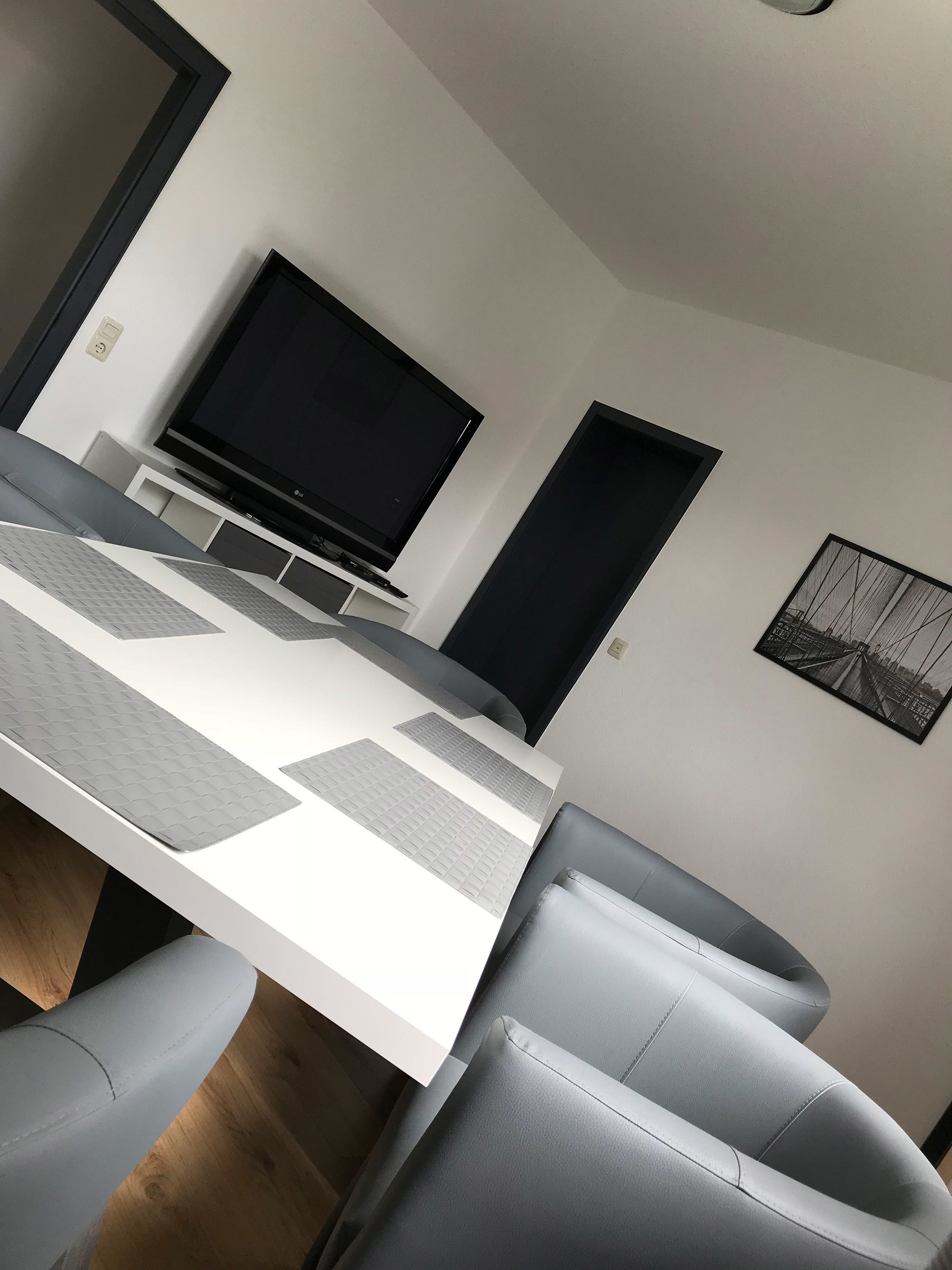 A modern dining area with a white table, grey cushioned chairs, a flat-screen TV, and a framed picture on the wall.