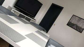 A modern dining area with a white table, grey cushioned chairs, a flat-screen TV, and a framed picture on the wall.