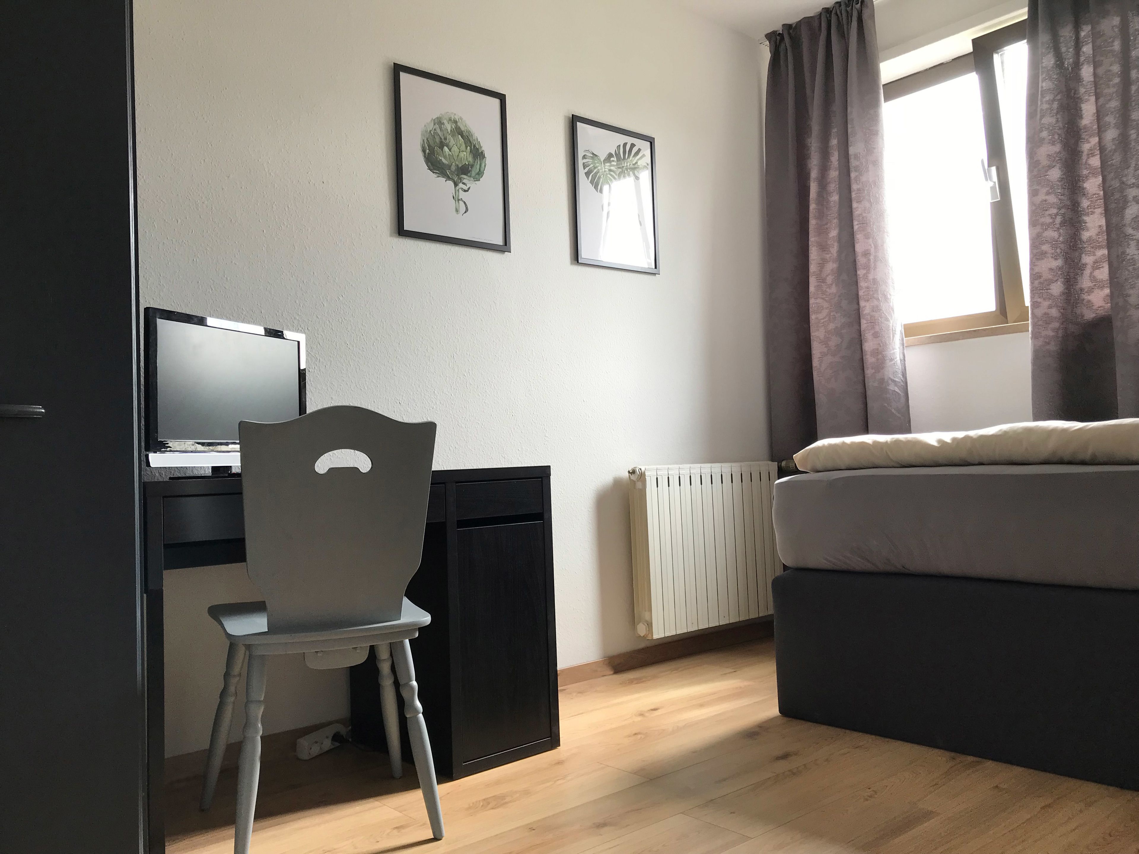 A bedroom with a bed, a desk with a computer, a chair, two framed pictures on the wall, a window with curtains, and a radiator.
