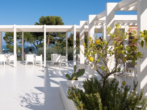 A white outdoor seating area with tables and chairs, surrounded by greenery and a view of the sea. - Hotel La Chiave dei Trabocchi (San Vito Chietino)